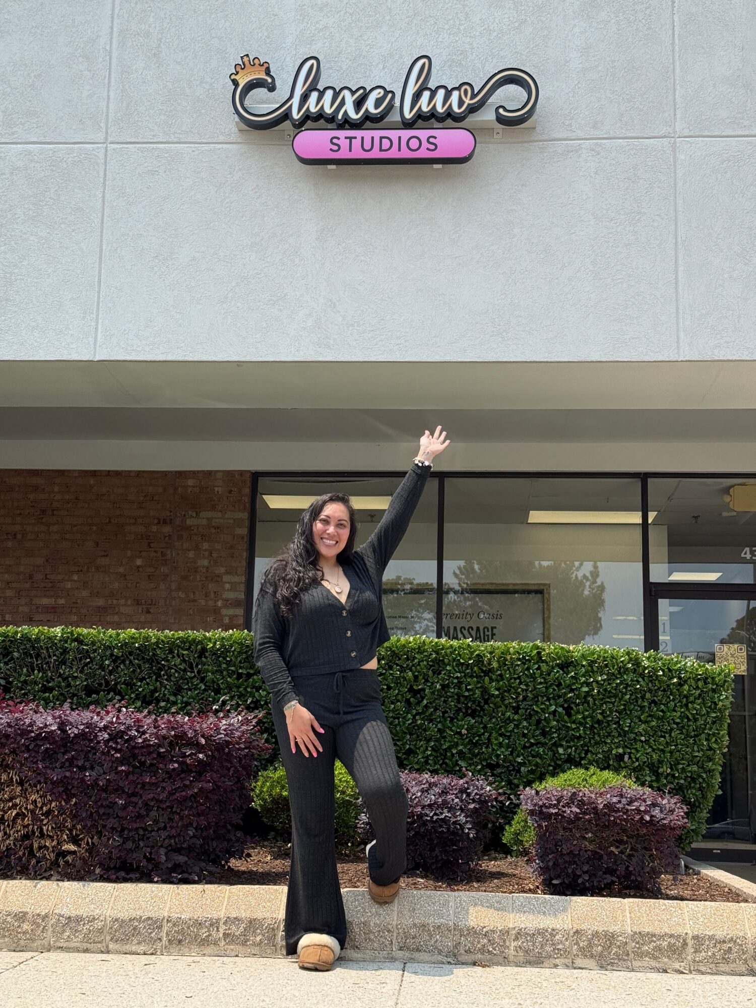 Woman standing outside a building, waving, with bushes and a sign reading 'Amore Studio' behind her.