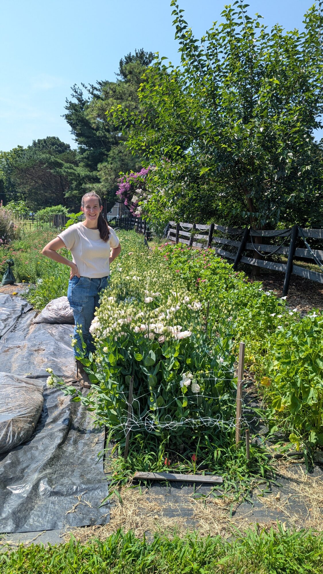 Woman standing in a garden with trees, plants, and a wooden fence, smiling with hands on hips.