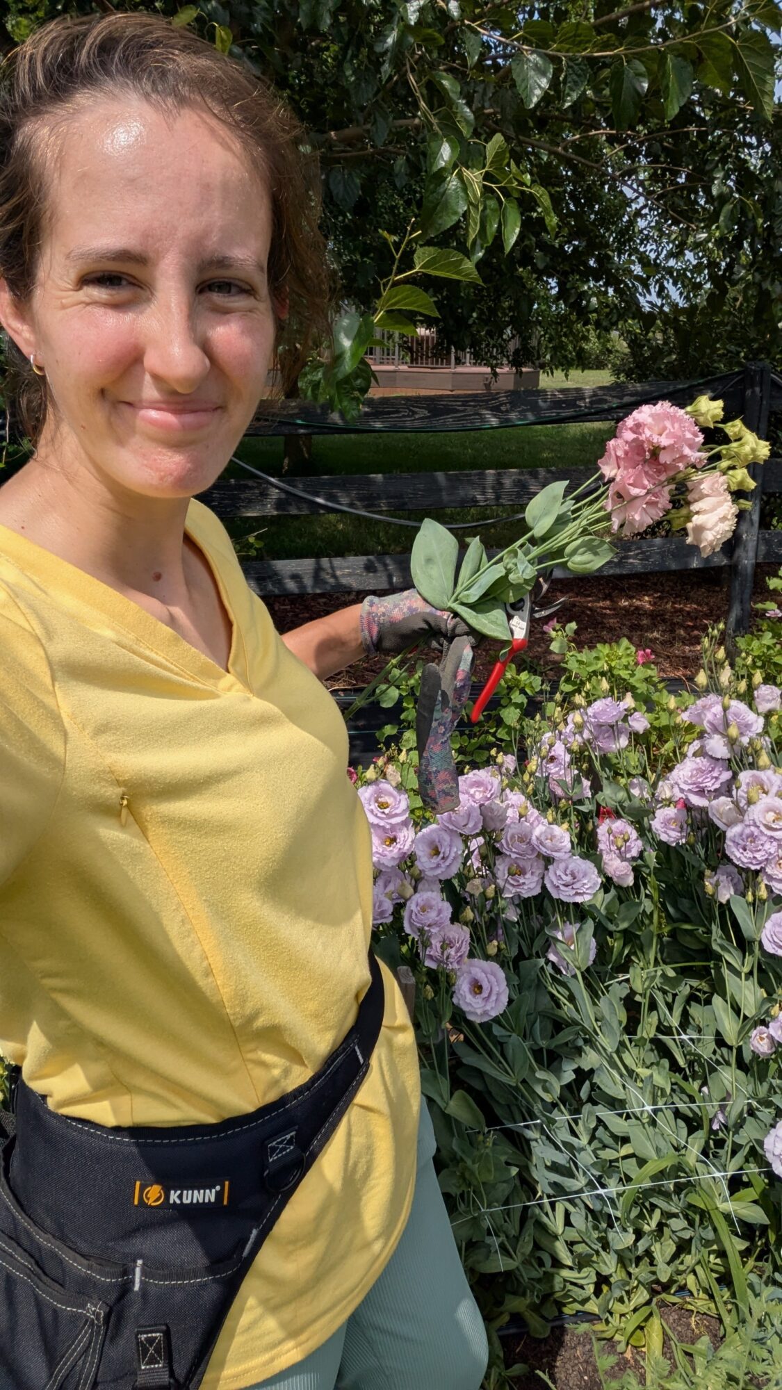 Woman in yellow shirt smiling outdoors with pink and purple flowers and green foliage behind her.