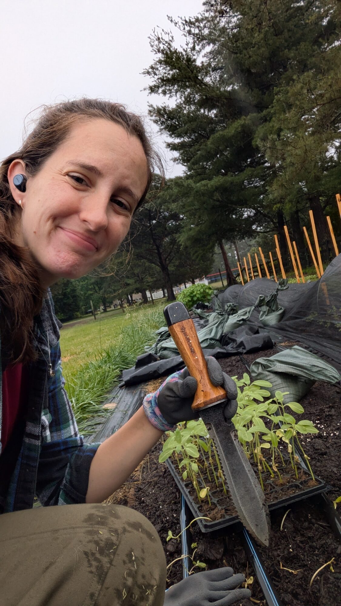 Young woman smiling outdoors, holding gardening tools, with plants and trees in the background.