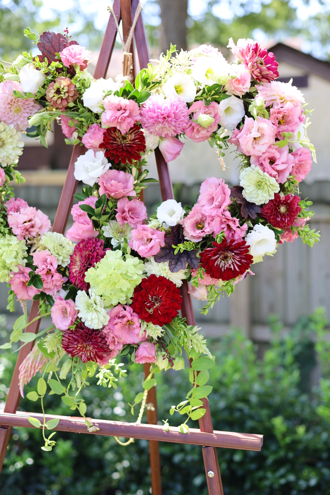 Pink, red, and white flowers arranged in a wreath on a pink metal stand outdoors with greenery in background