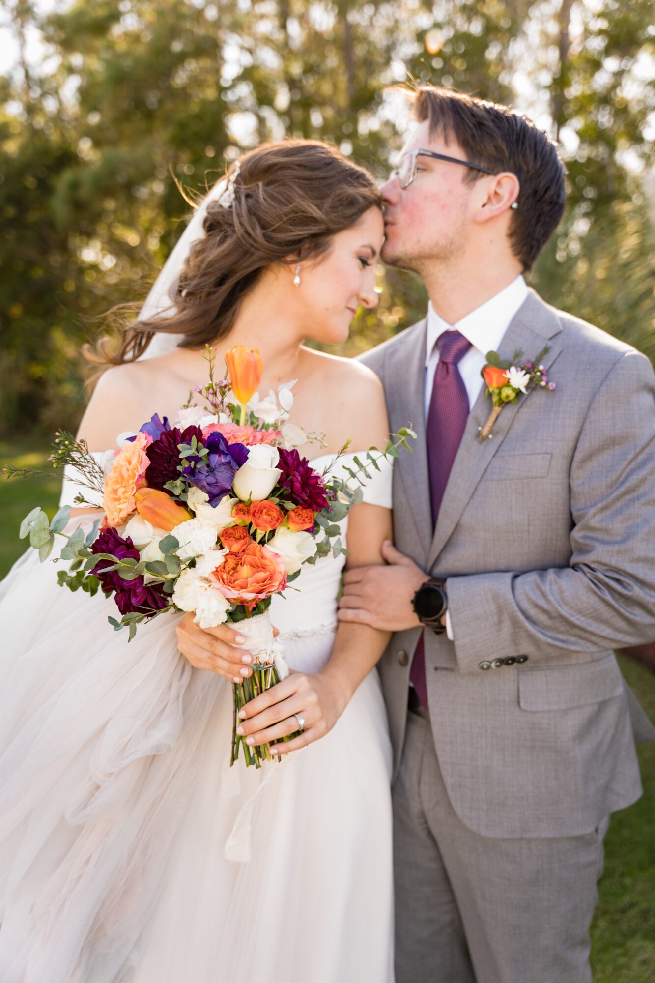 A bride and groom share a kiss outdoors, with trees in the background. The bride holds a colorful bouquet, and the groom wears a gray suit.