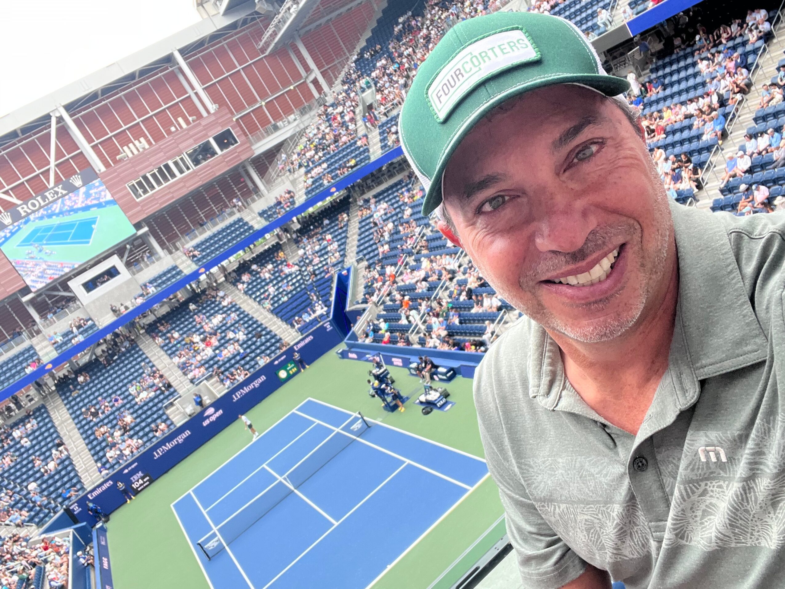 Smiling man taking a selfie at a tennis stadium with a court and spectators in the background.