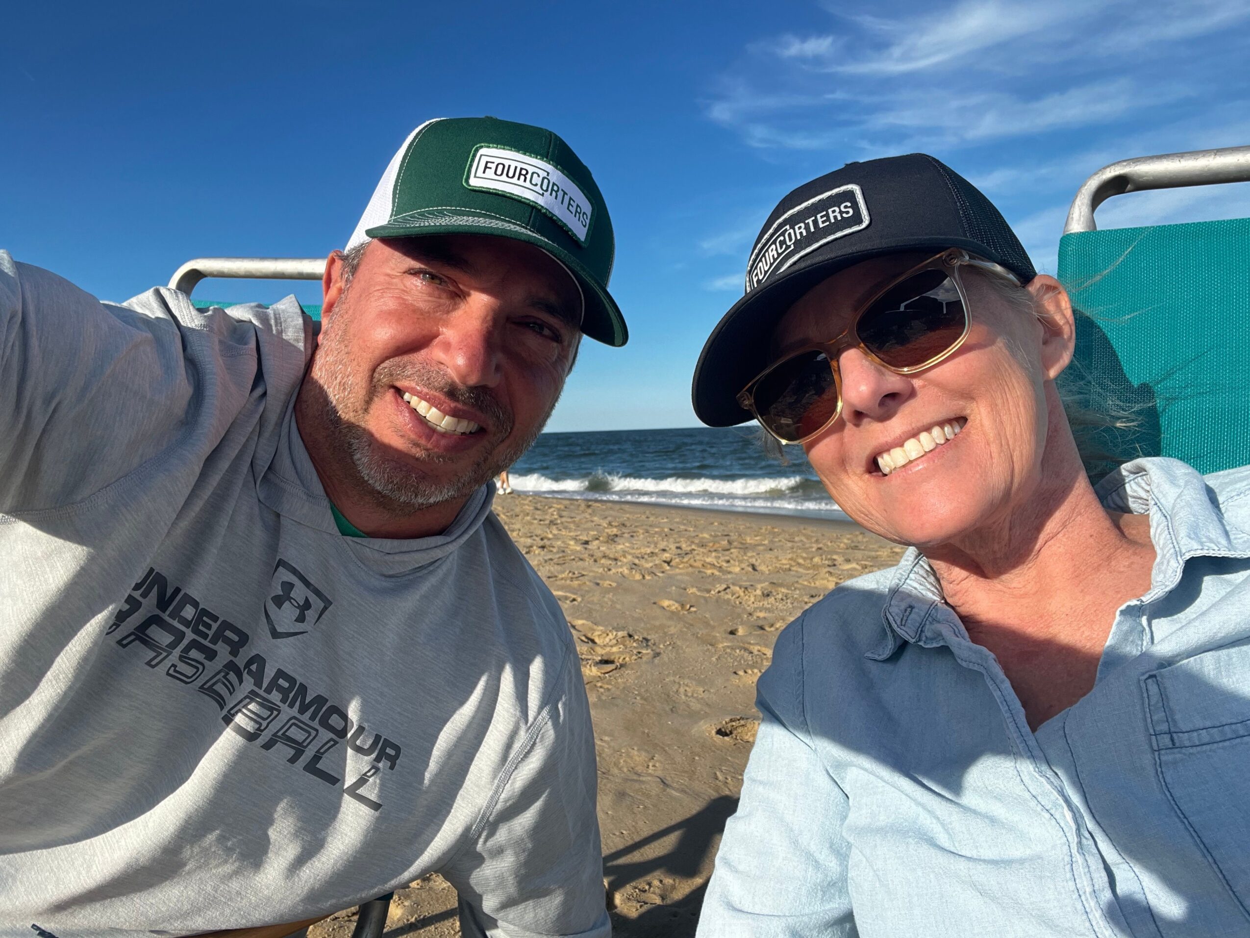 Two people smiling, wearing hats and sunglasses, lying on a beach with blue sky and sand visible.