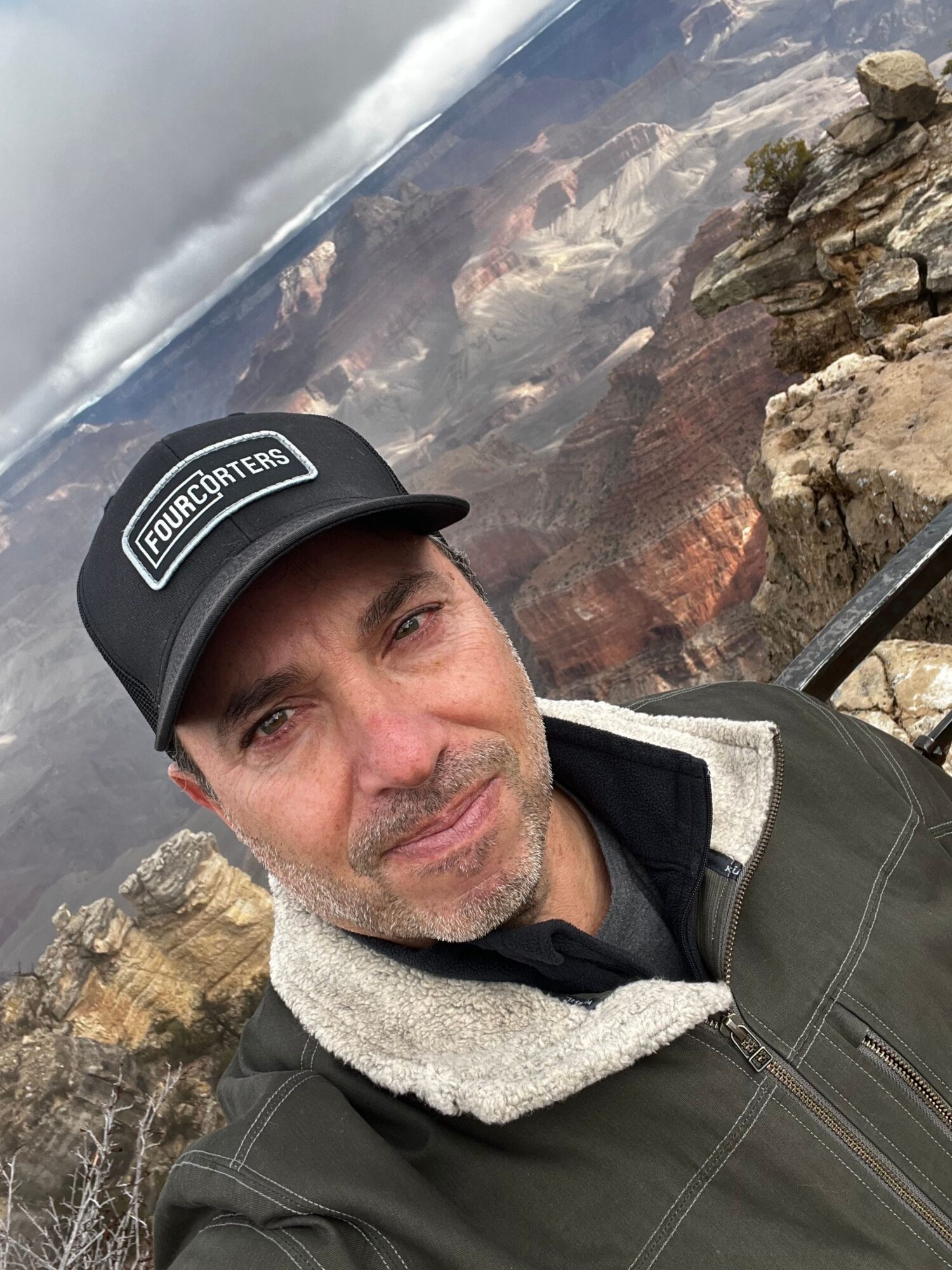 Man wearing a cap and jacket taking a selfie outdoors with rocky landscape and cloudy sky in background.