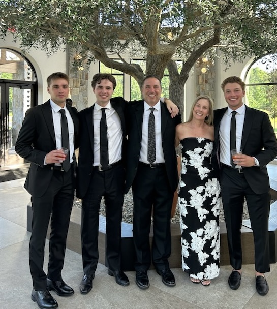 Group of five people dressed in formal attire standing outdoors under a tree, smiling, with two holding drinks.
