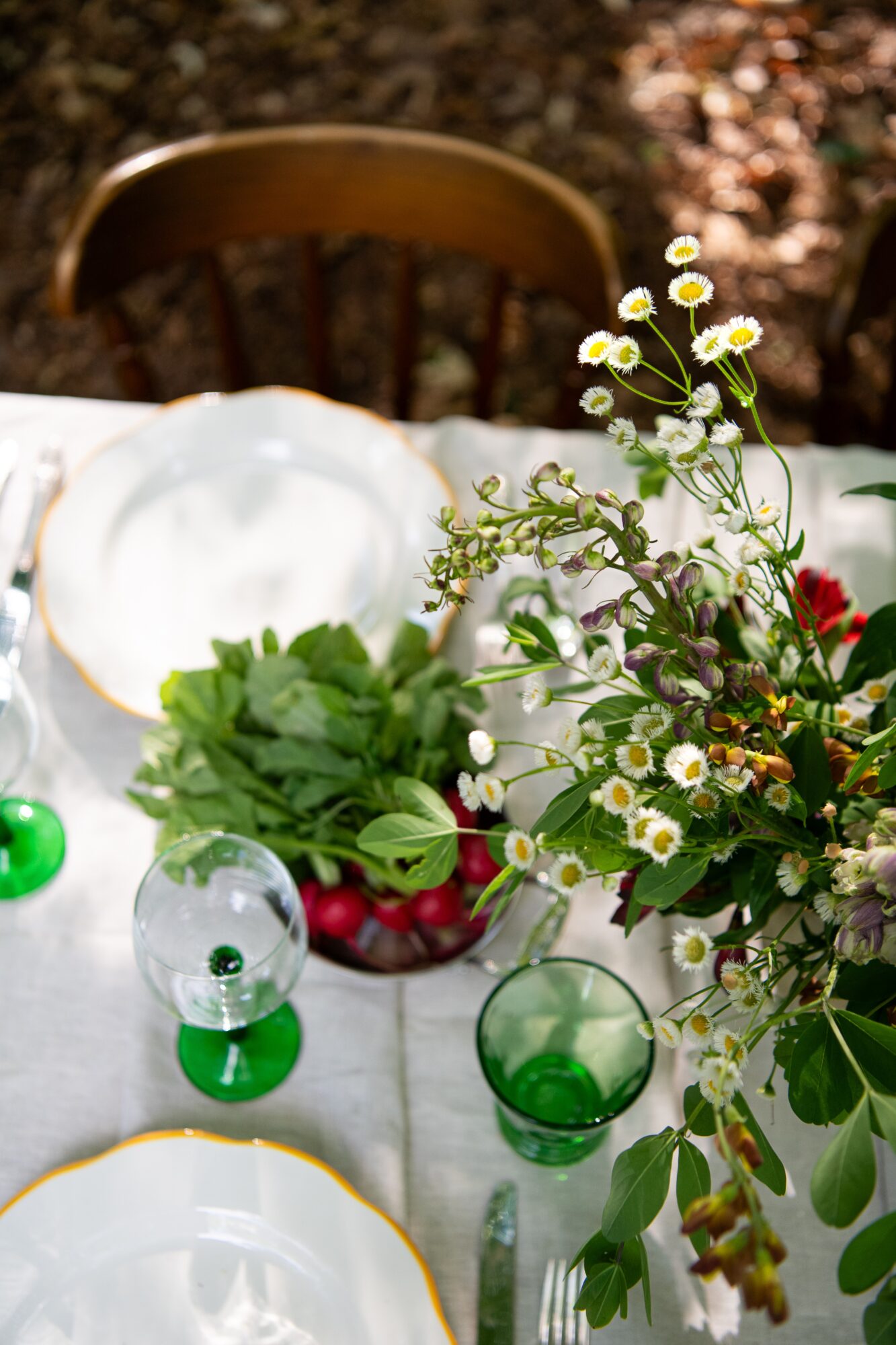 Table with a floral arrangement, green glasses, and a bowl of cherries, with a wooden chair in the background.