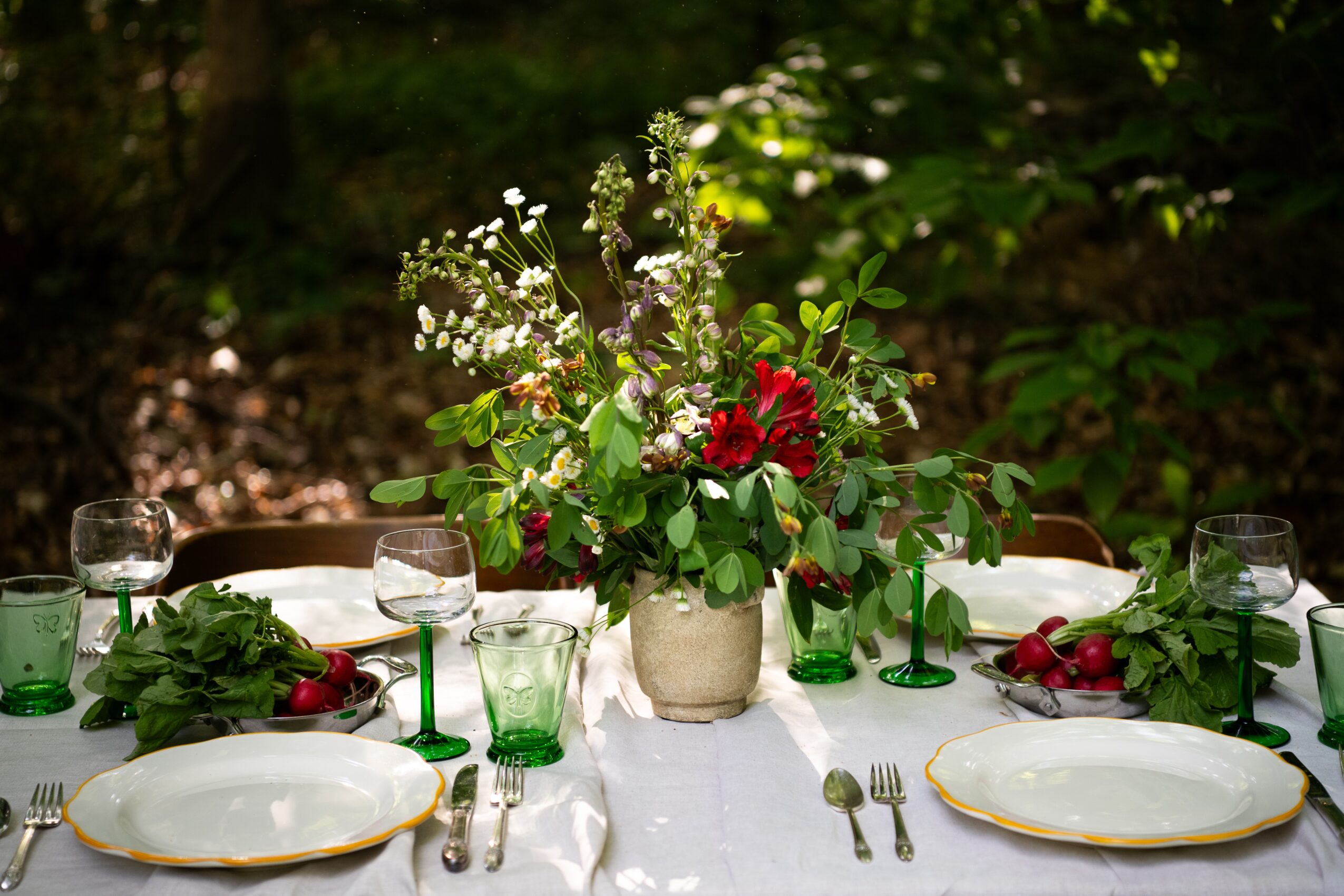 Table set outdoors with a floral centerpiece, plates, glasses, and utensils, surrounded by greenery.