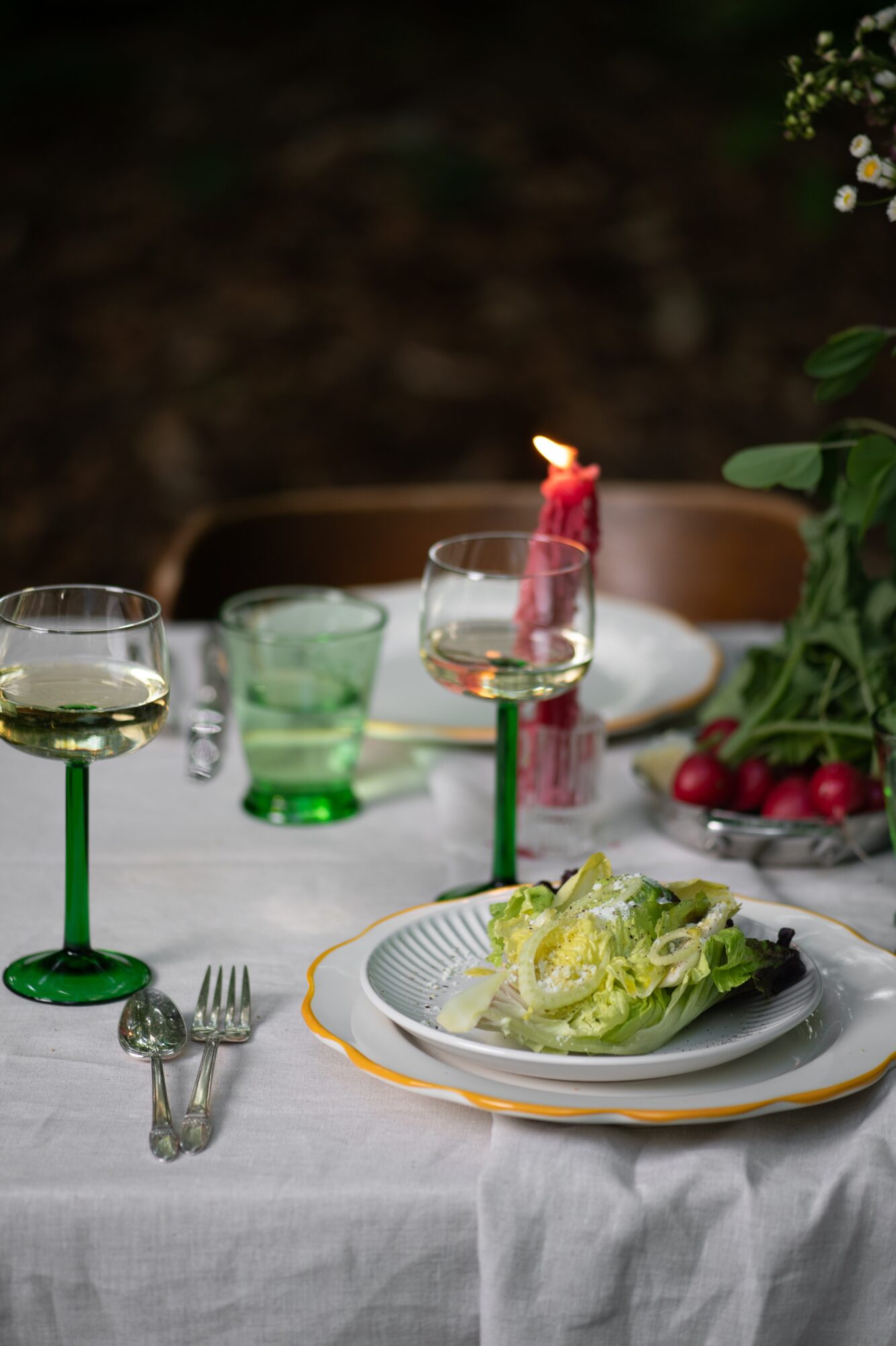 Table with wine glasses, a plate of salad, a lit candle, and a bowl of cherries, set for a meal.
