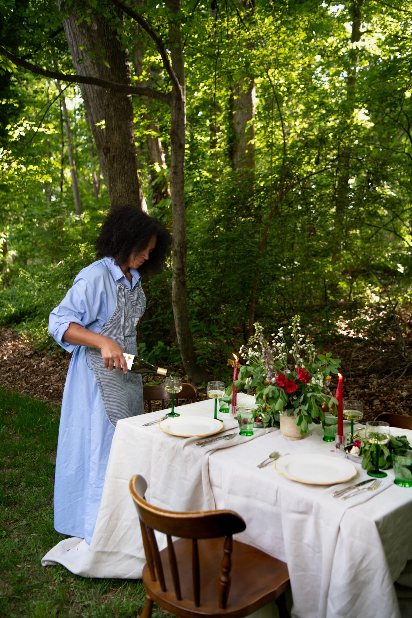 Person in a blue dress setting a table outdoors with flowers and candles, surrounded by green trees.