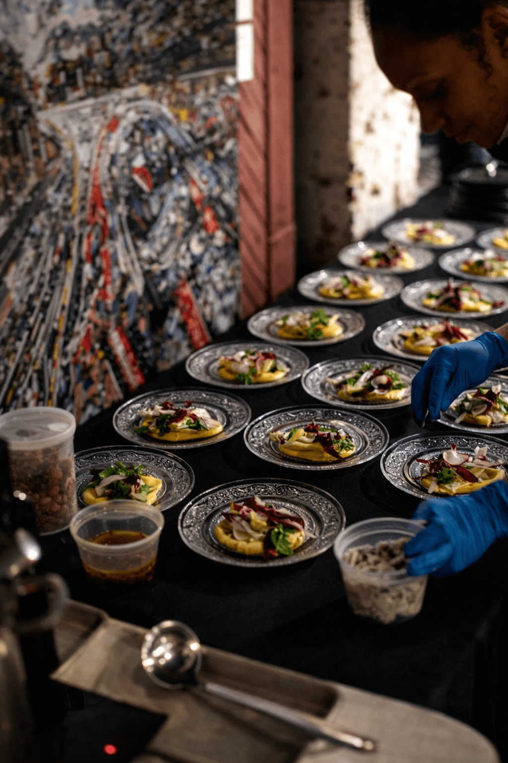Person wearing blue gloves preparing multiple plates of food on a black table, with a colorful mosaic background.