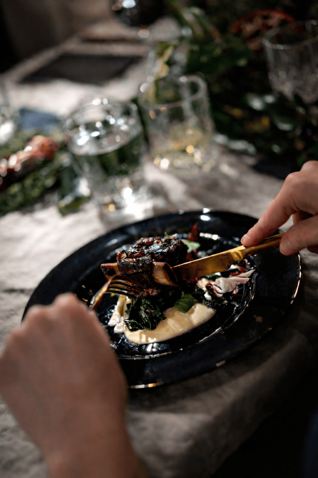 Person cutting a dessert on a black plate with a fork, with glasses and a tablecloth in the background.