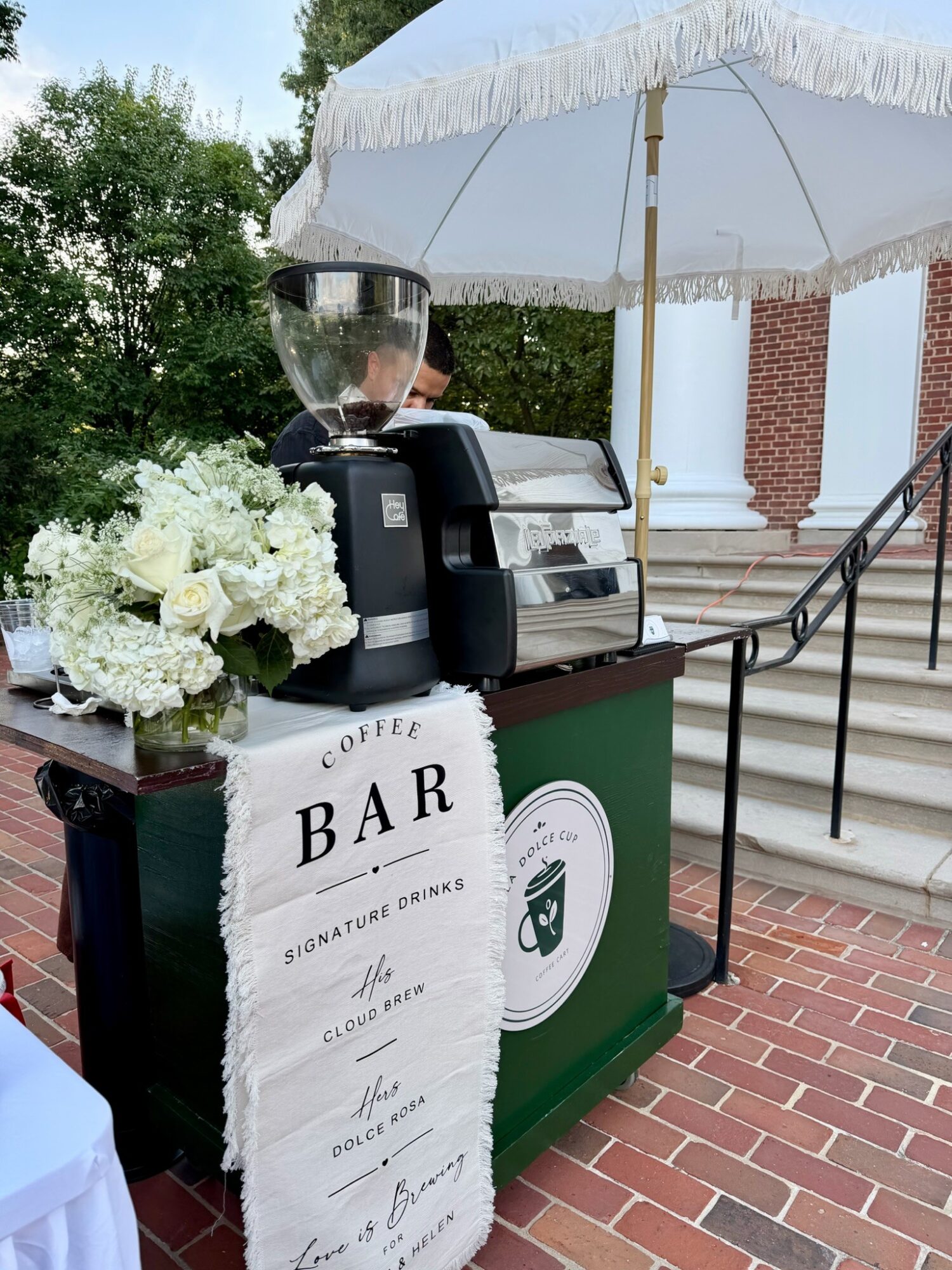 Coffee bar setup outdoors with flowers, umbrella, and stairs in background, featuring a sign and equipment.