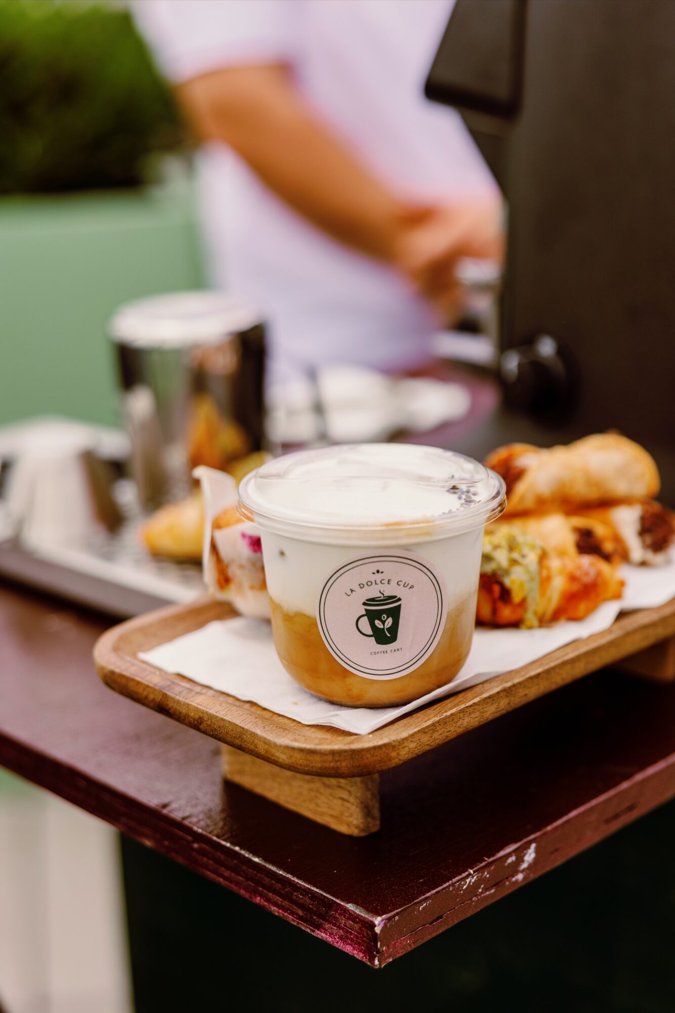 Cup of coffee with a logo on a wooden tray with food, person in background, blurred, indoors.