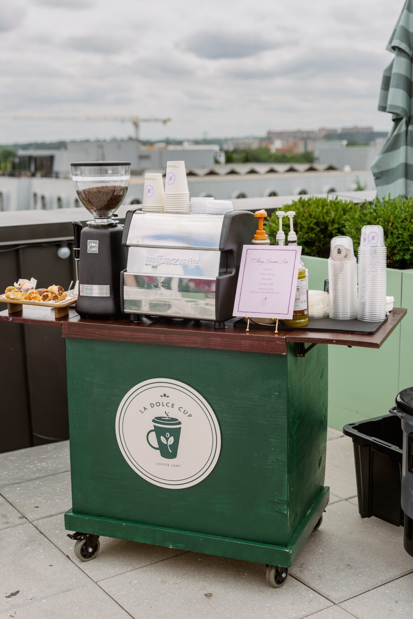Coffee station with a coffee grinder, cups, and a menu on a green cart outdoors, city skyline in background.