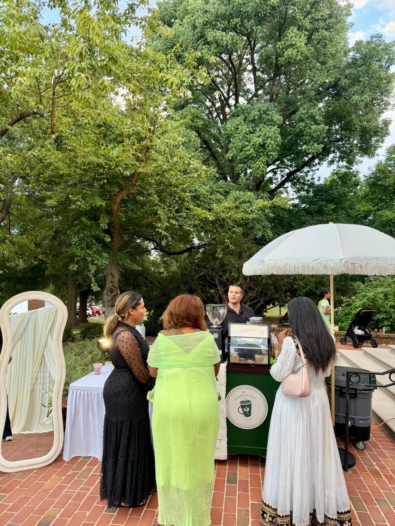 Four women stand outdoors near a table with a laptop and a white umbrella, surrounded by trees and park benches.