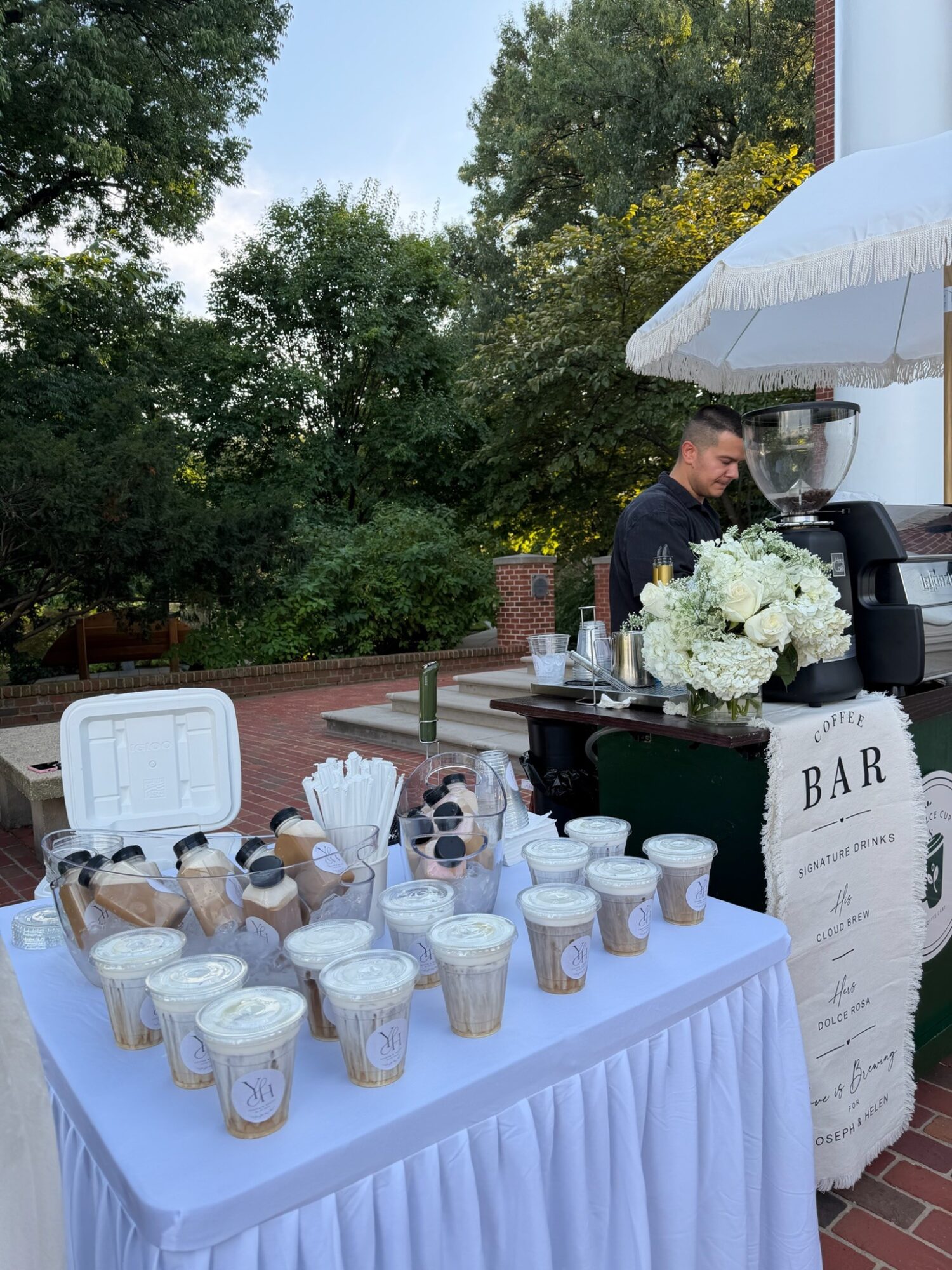 Table with drinks and a bar setup outdoors, person preparing beverages, surrounded by trees and a white umbrella.