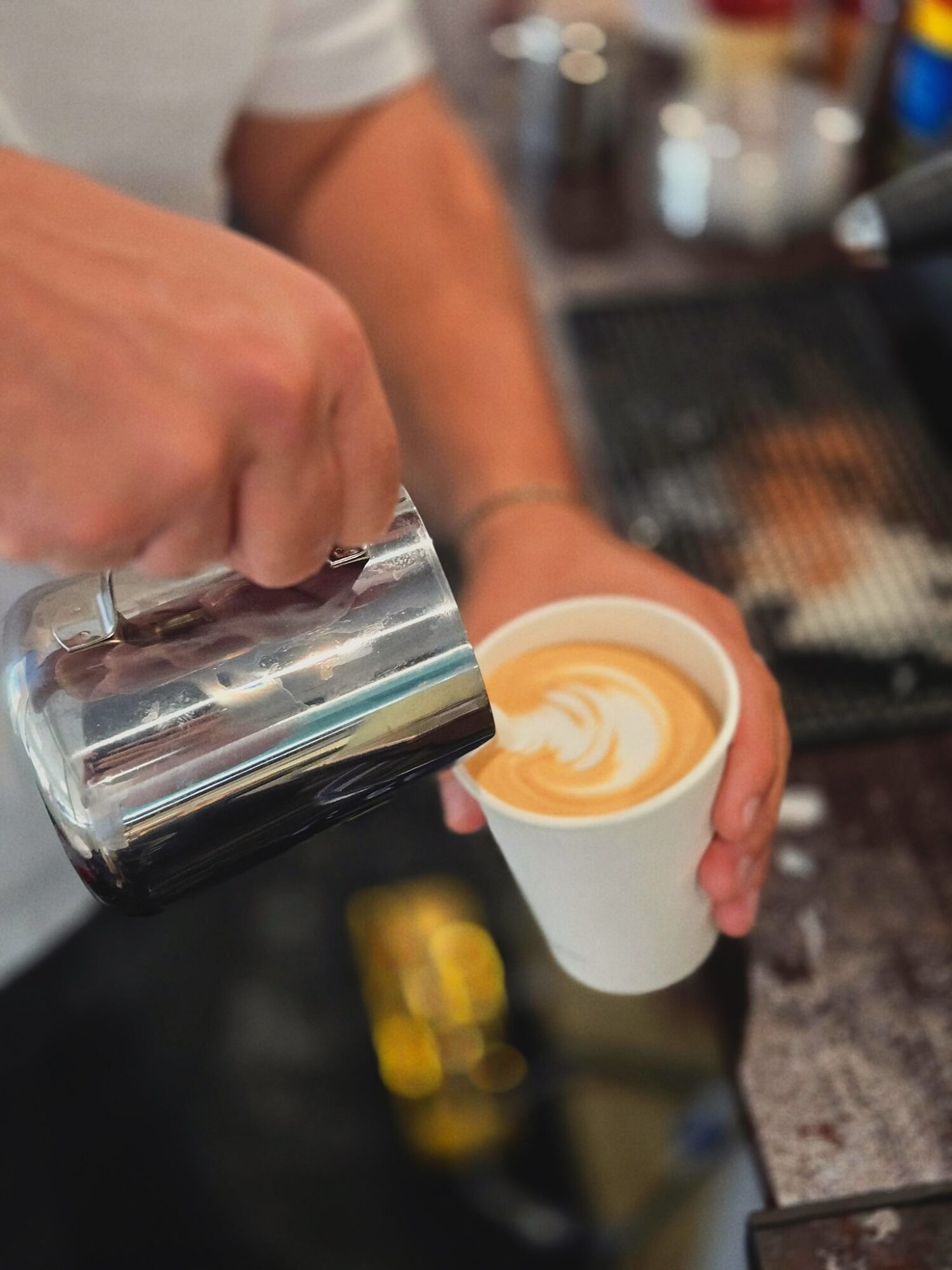Person pouring steamed milk into a cup of coffee, creating latte art, in a cafe setting.