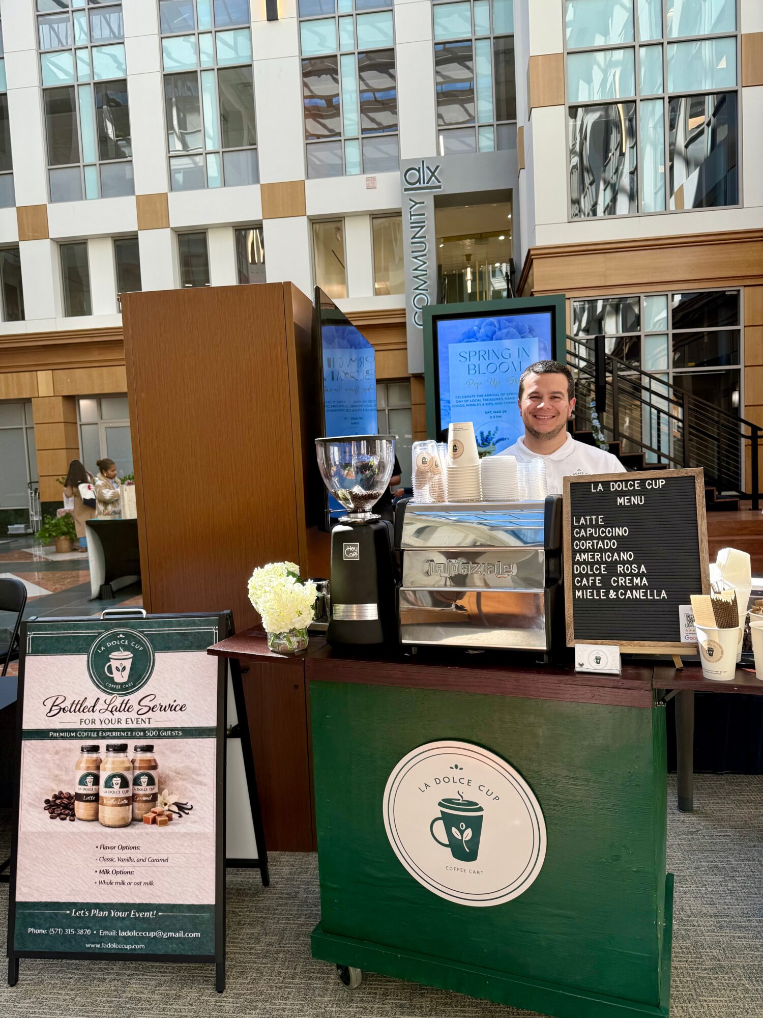 Coffee stand with a smiling barista, menu board, and promotional sign outside a modern building.