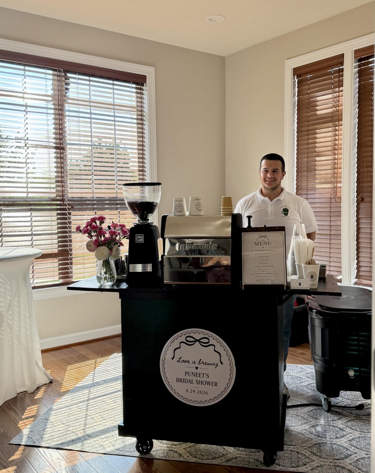 Person standing behind a coffee cart with a coffee grinder, cups, and flowers in a bright room with large windows and wooden blinds.