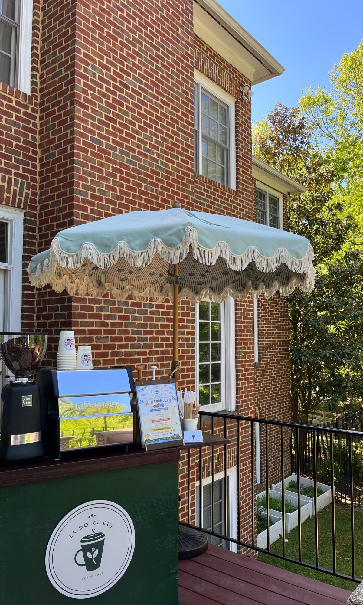 Outdoor cafe setup with a white scalloped umbrella, a coffee machine, and a menu on a green counter, next to a brick building.