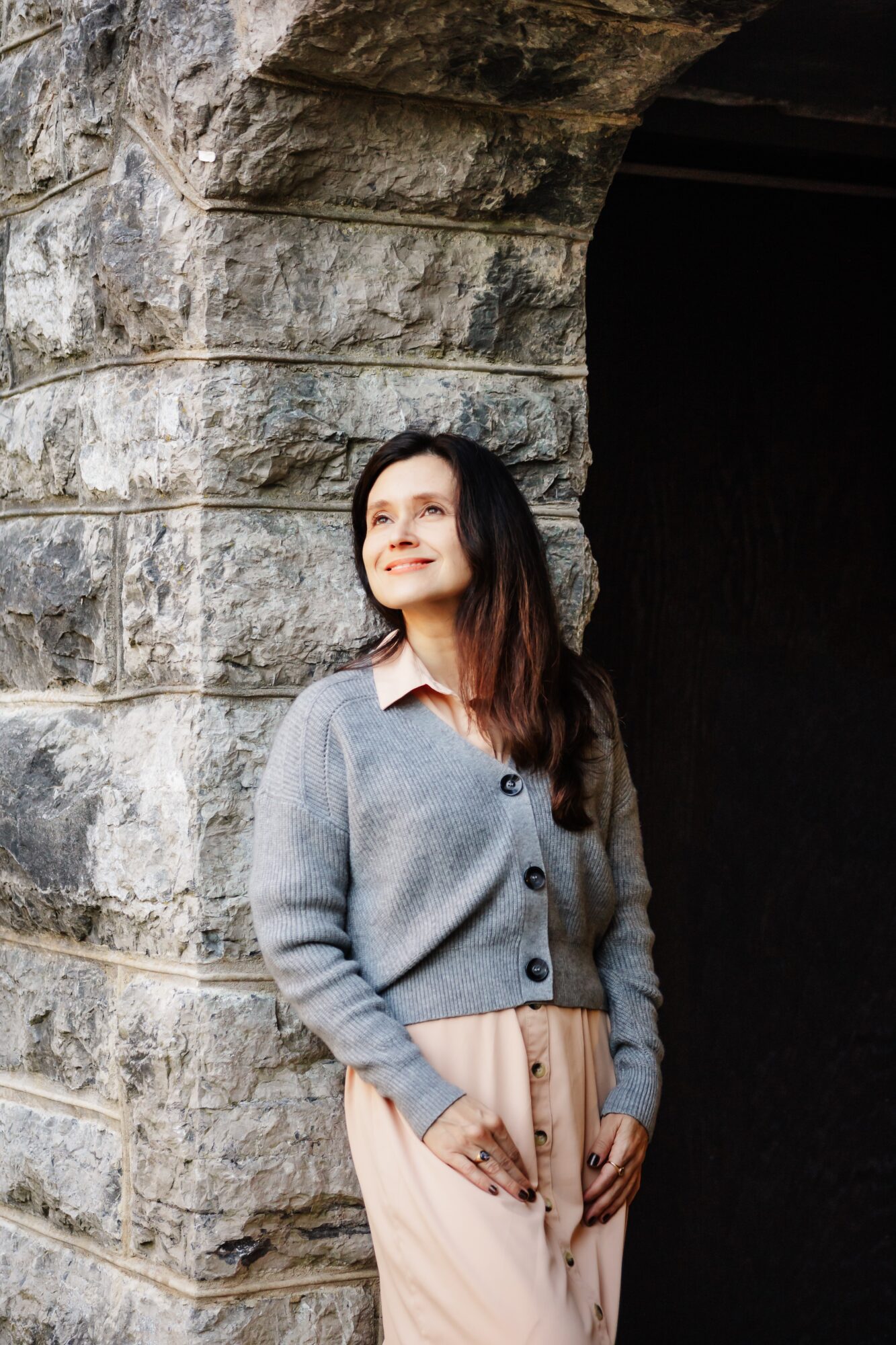 Woman with long dark hair wearing a gray cardigan and beige skirt standing near a stone archway, smiling and looking upward.