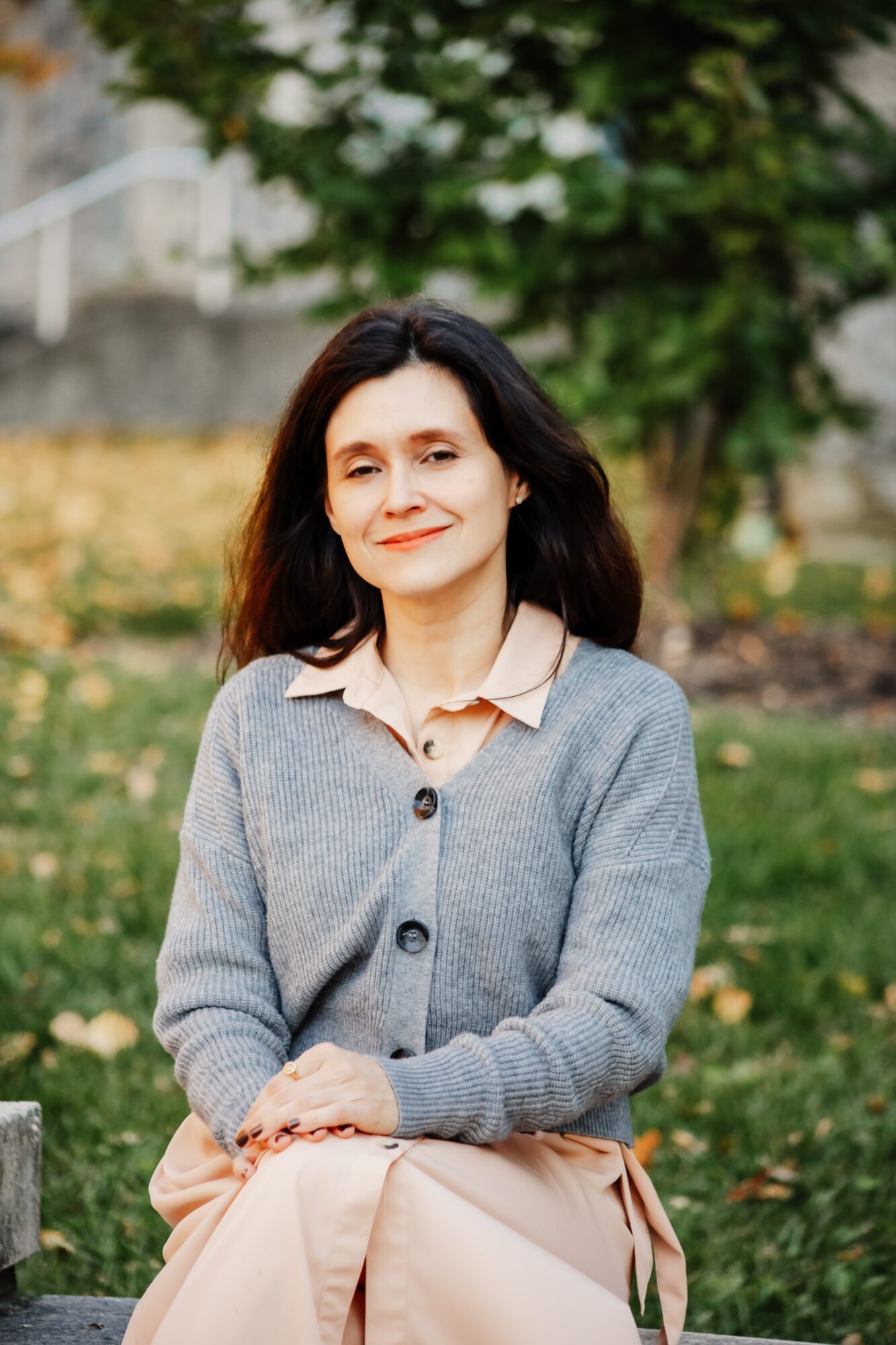 Woman with dark hair sitting outdoors in a park, wearing a gray cardigan and light-colored blouse, smiling.