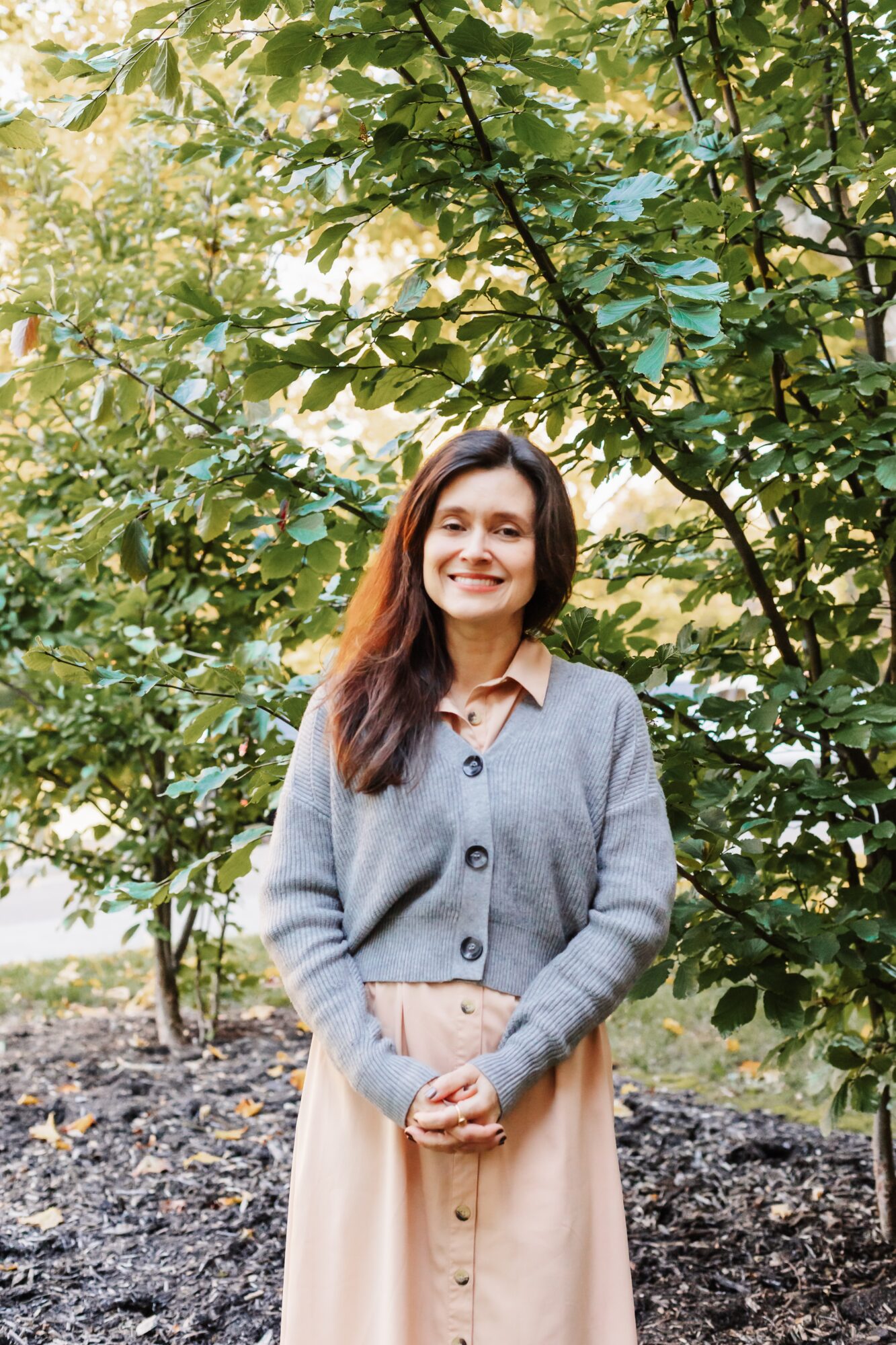 Young woman with long dark hair smiling outdoors, wearing a gray cardigan and beige skirt, standing among green trees.