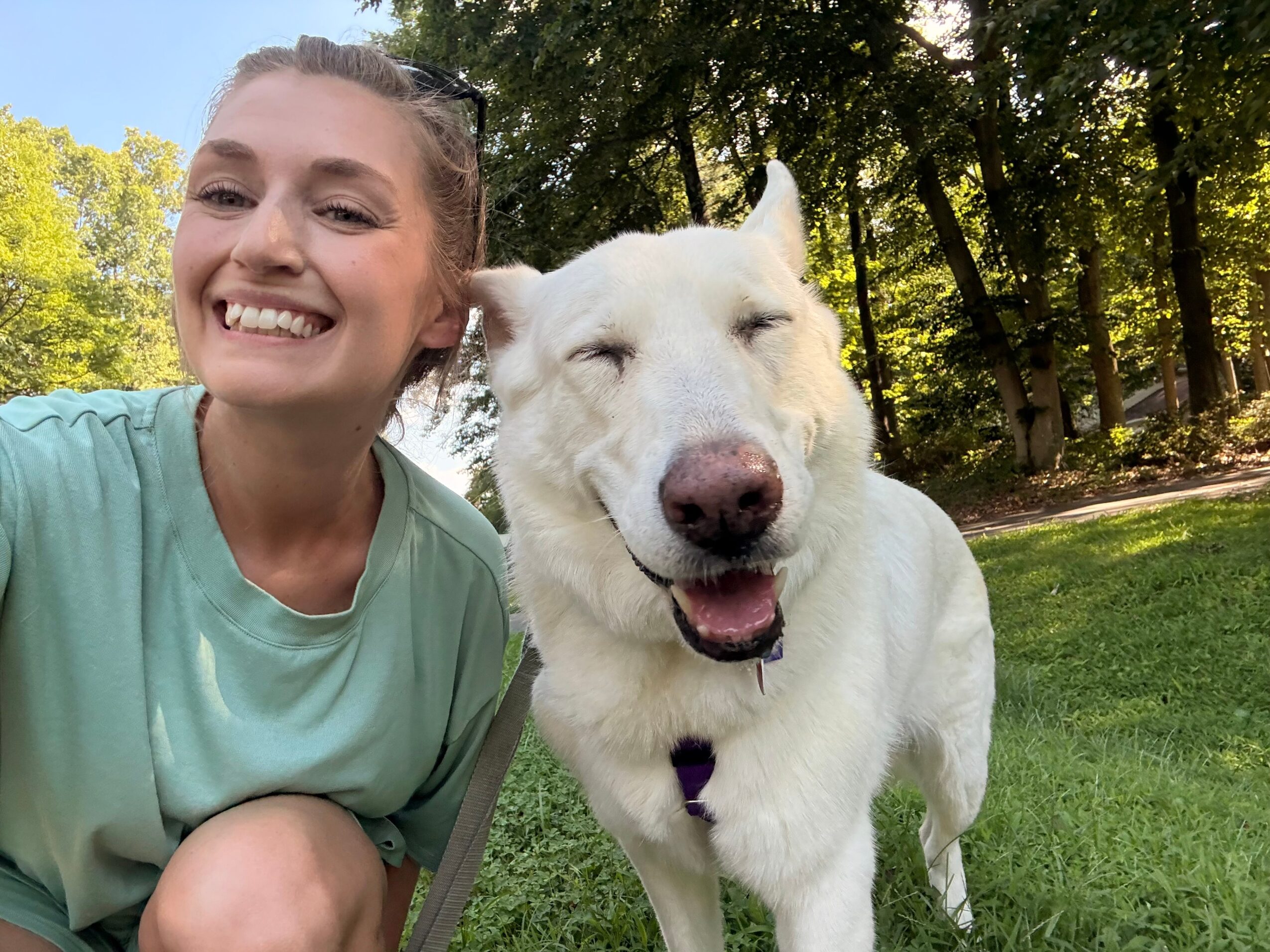 Smiling woman with a white dog outdoors in a park, trees and grass in background.