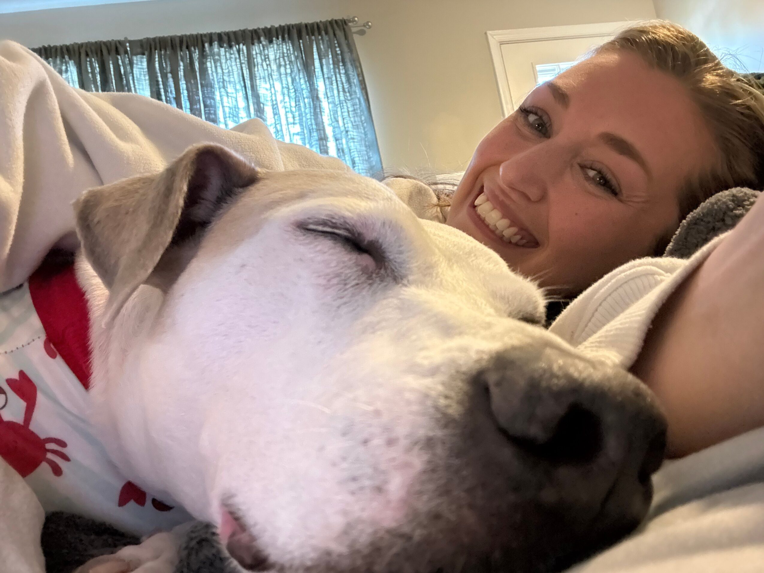 Person smiling with a dog resting its head on their shoulder, both lying down indoors.