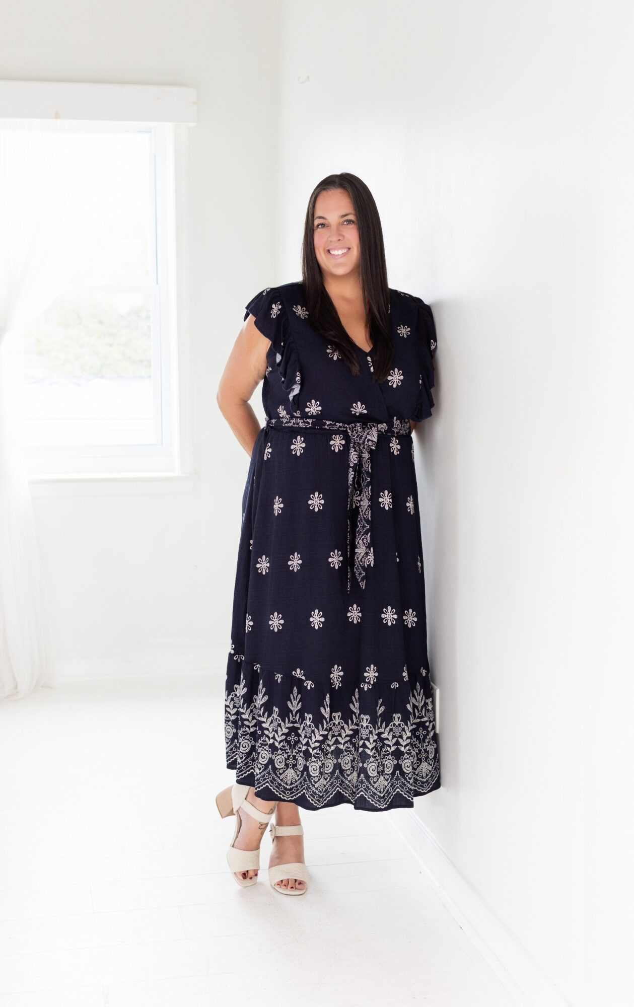 Woman in a navy dress with white patterns, standing in a bright room near a window, smiling.