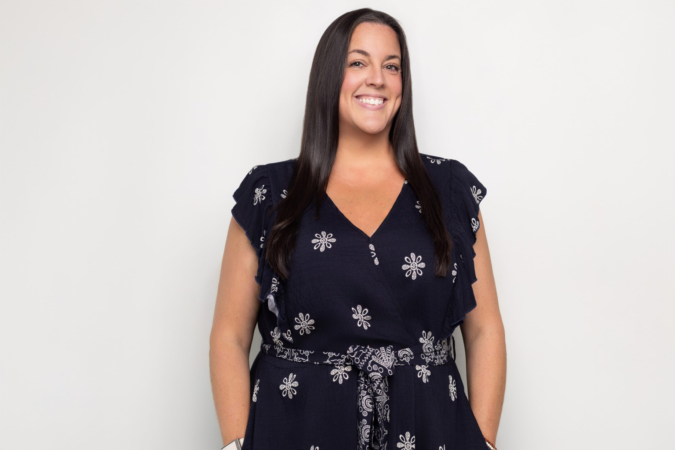 Smiling woman with long dark hair wearing a navy dress with floral patterns, standing against a plain white background.