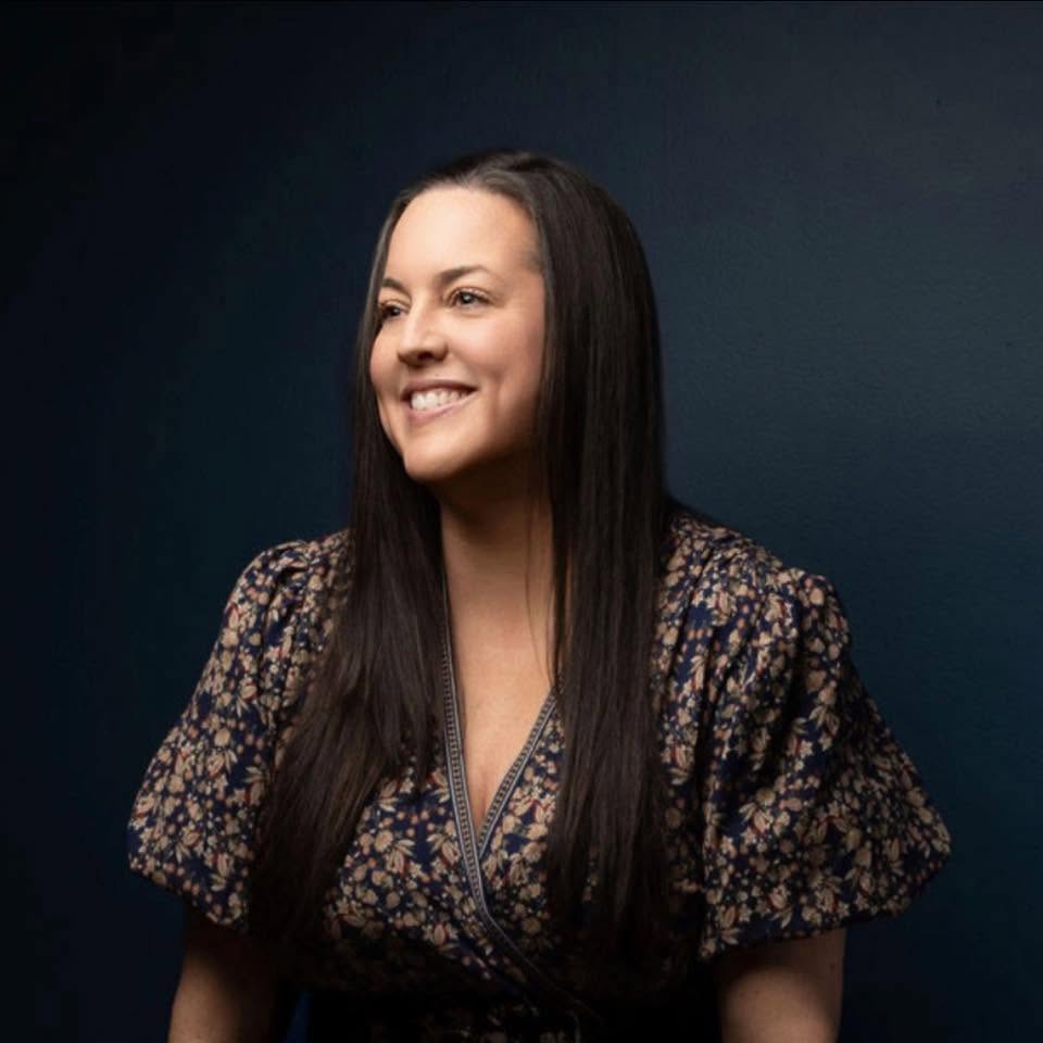 Smiling woman with long dark hair wearing a patterned blouse against a dark background.