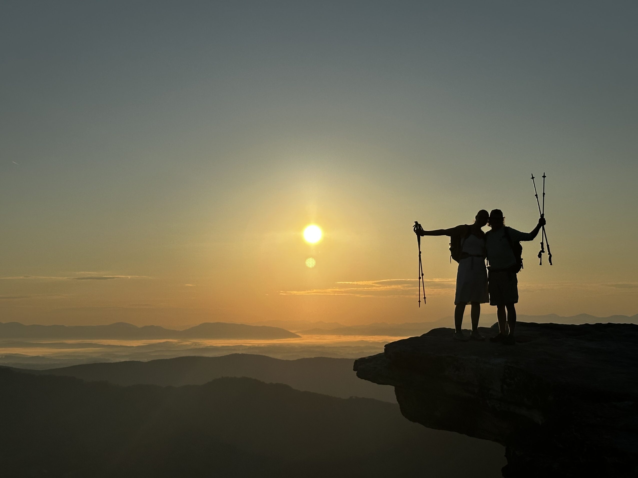 Two people stand on a cliff edge holding a bow and arrow, watching a sunset over mountains.