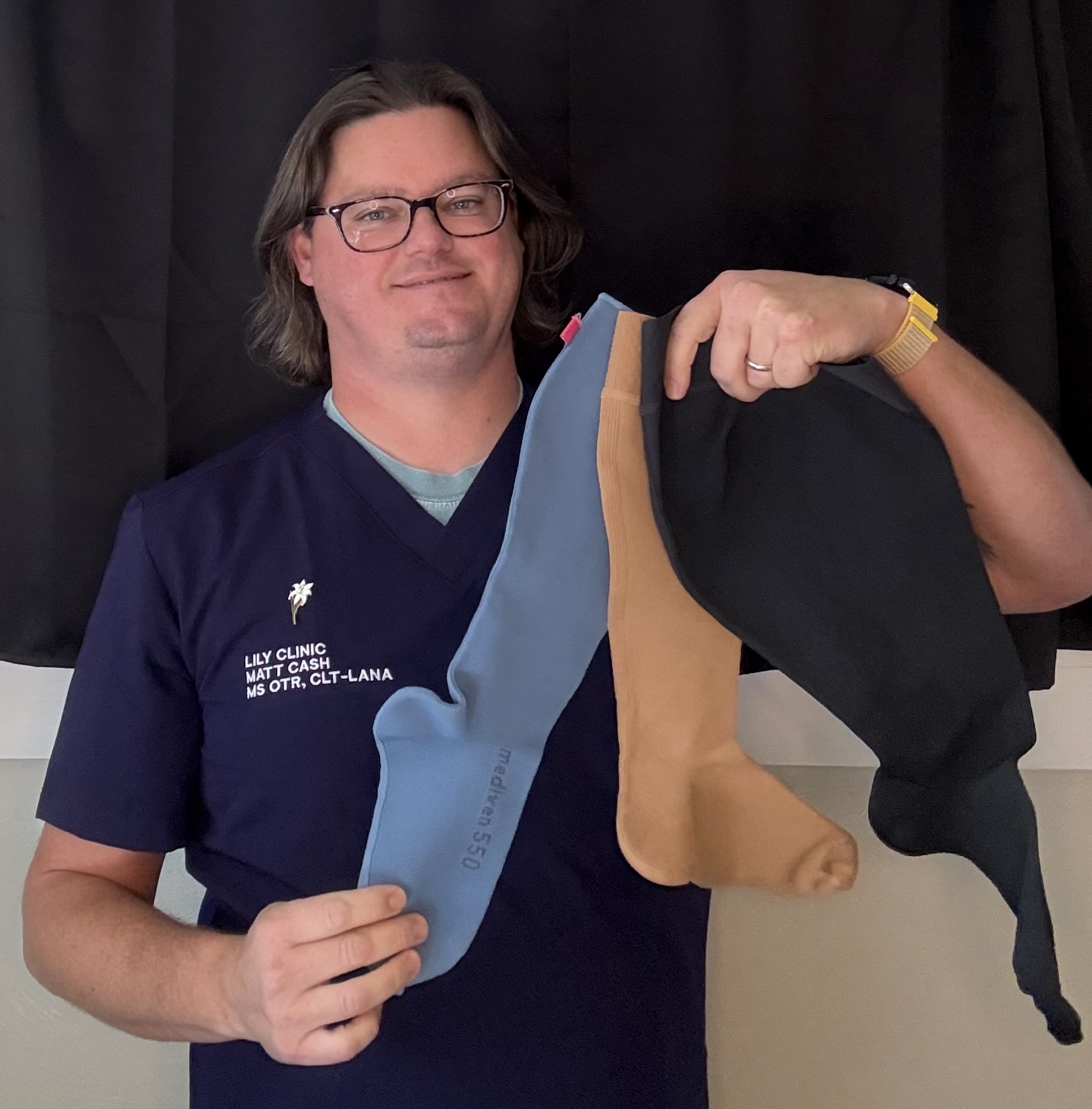 Person with glasses and shoulder-length hair holding three fabric items, wearing a navy blue shirt with text, standing in front of a black background.