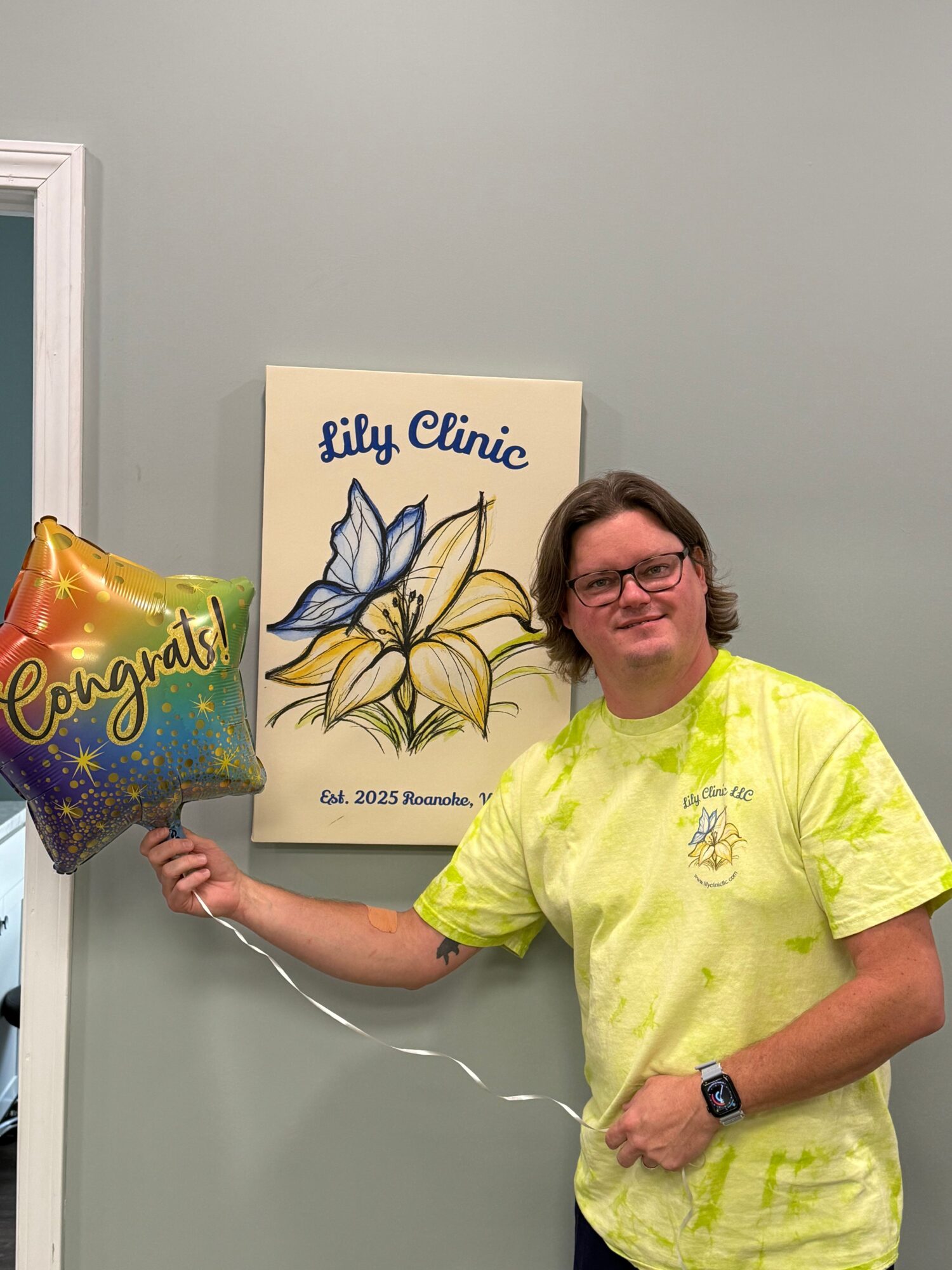 Woman in yellow shirt holding a star-shaped balloon, standing next to a sign with a flower illustration, in a room with gray walls.