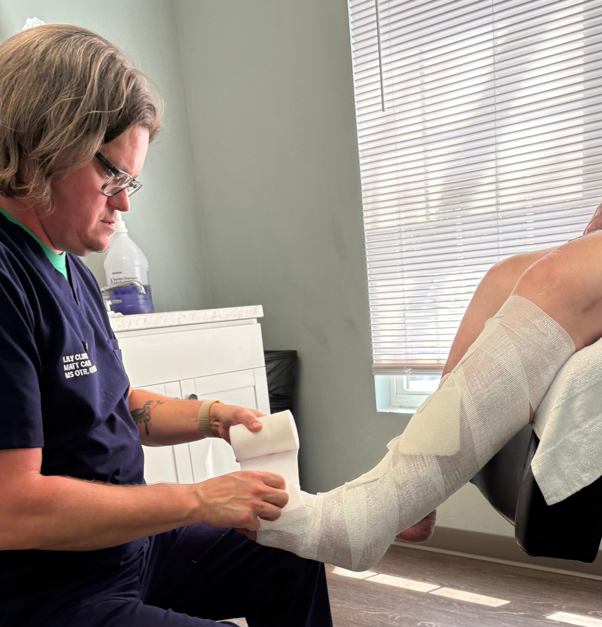 Medical professional examining a patient's leg in a clinical setting with blinds on the window.