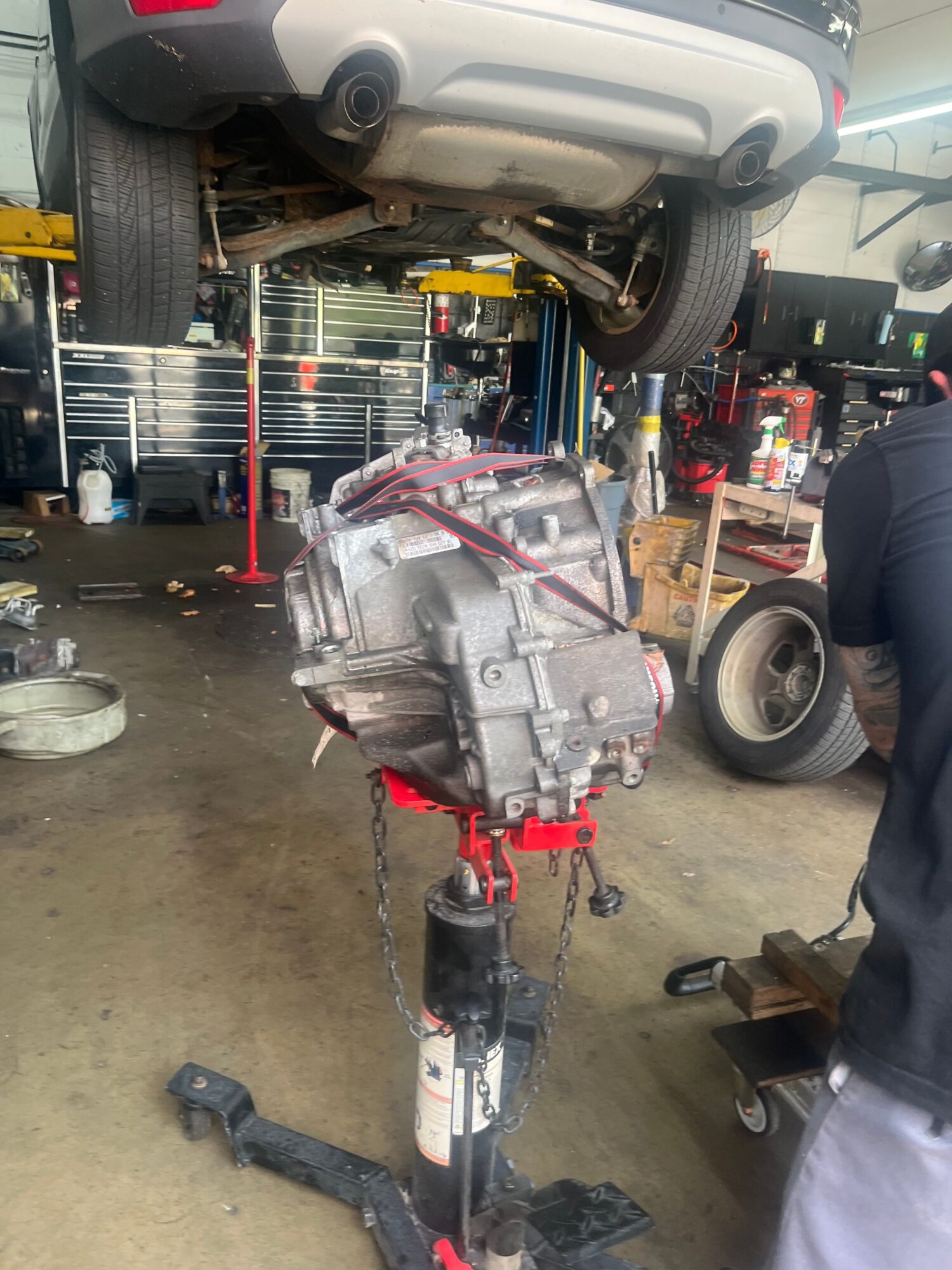 Car lifted with engine removed, workshop with tools and equipment, person working nearby, tires and parts visible.