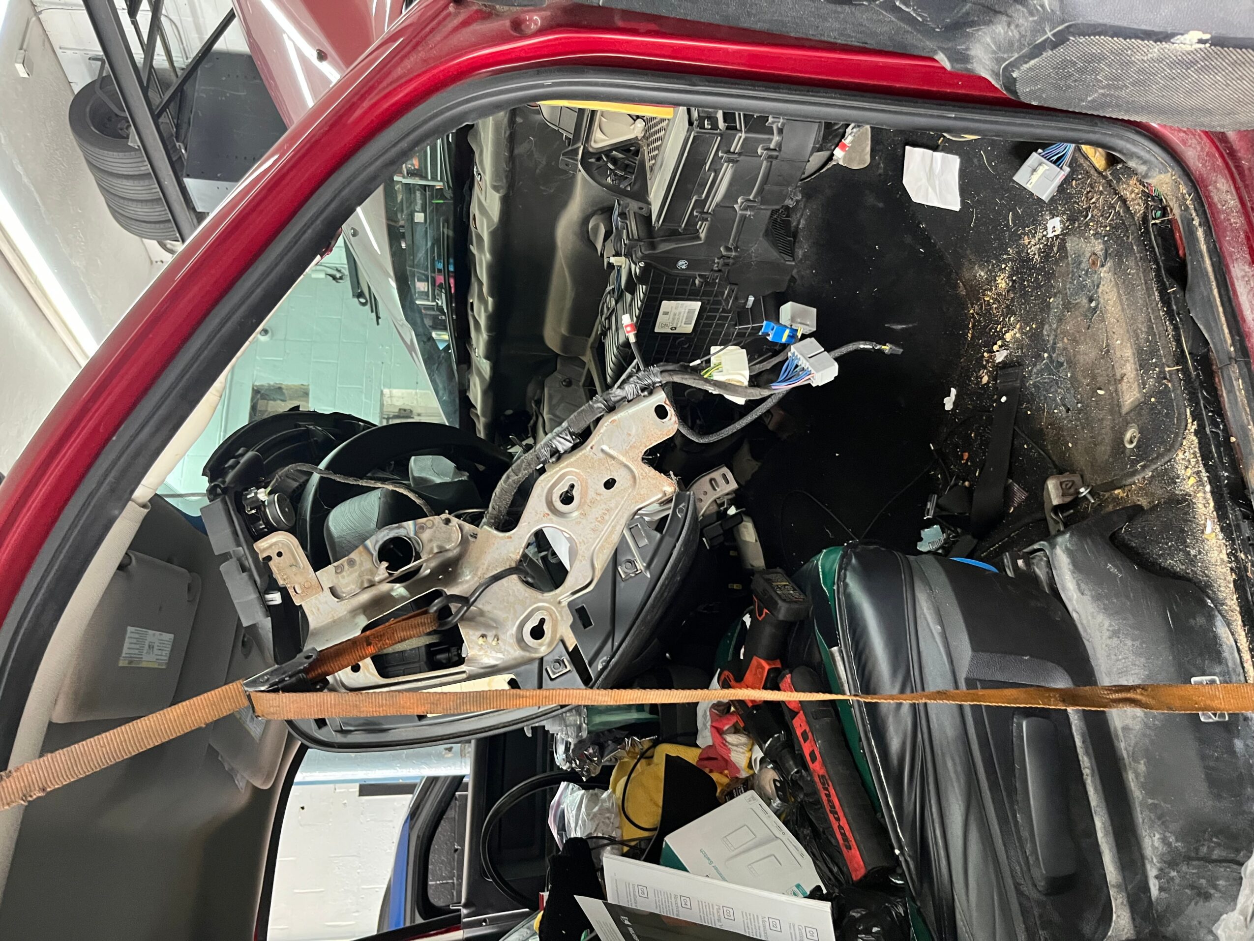 View of a vehicle's interior with the dashboard removed, exposing steering column and wiring, with a strap across the image.