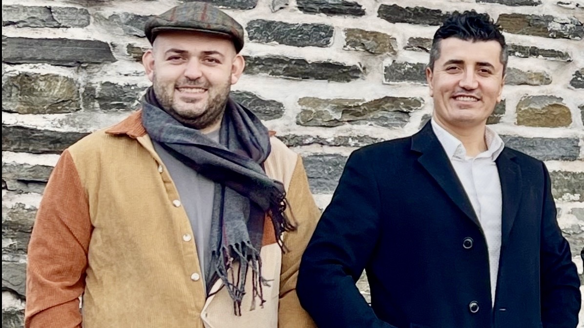 Two men standing outdoors in front of a stone wall, smiling, dressed in casual and formal clothing.