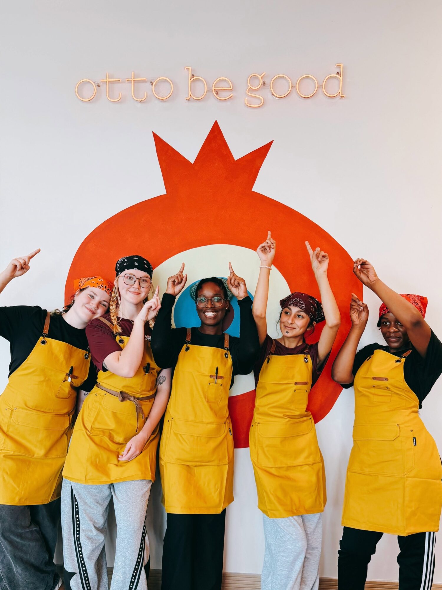 Group of five people wearing yellow aprons, standing in front of a wall with a large red star and text, smiling and raising hands.