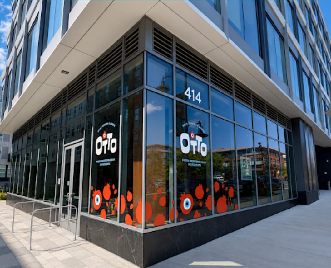 Corner of a modern glass building with the number 414 and a logo with orange circles, reflecting trees and sky.