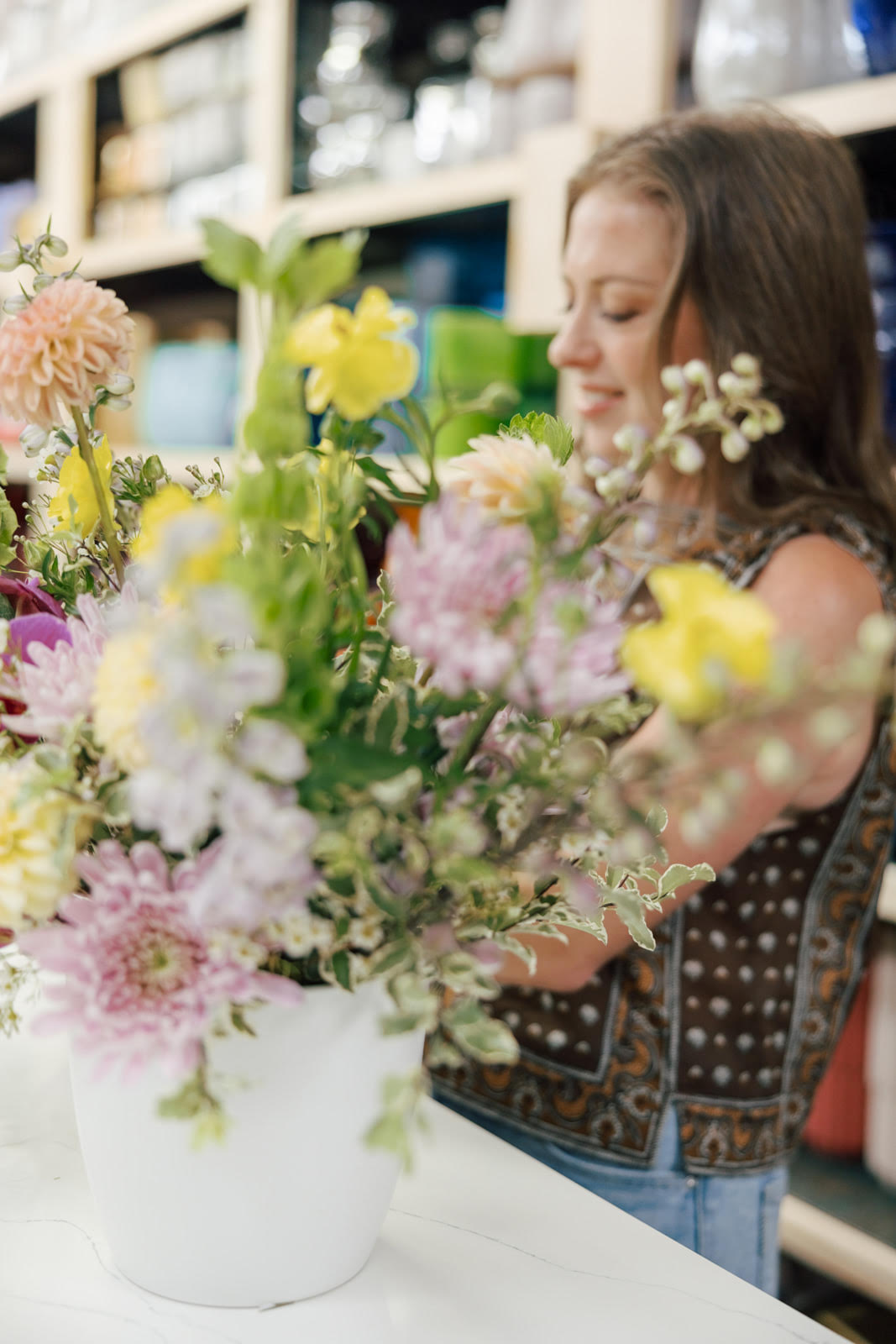 Woman smiling at a large colorful flower arrangement in a white vase, in a store setting.