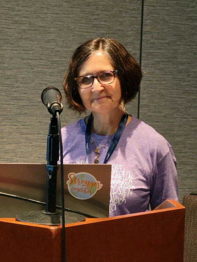 Woman with glasses and short brown hair standing behind a podium, wearing a purple top and a name badge.