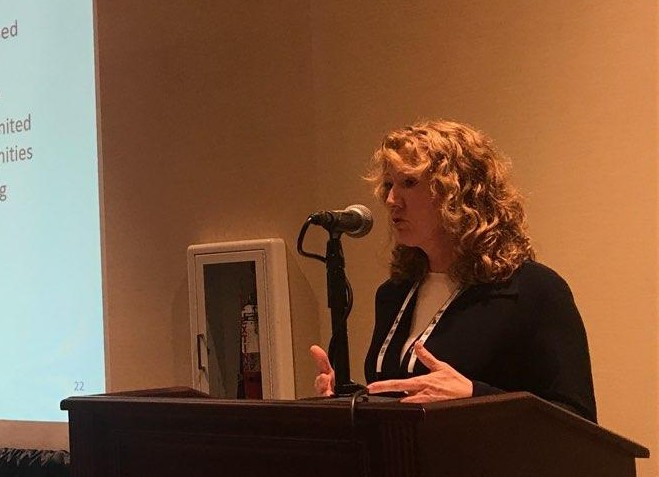 Woman with curly hair speaking at a podium with a microphone, in a room with beige walls.