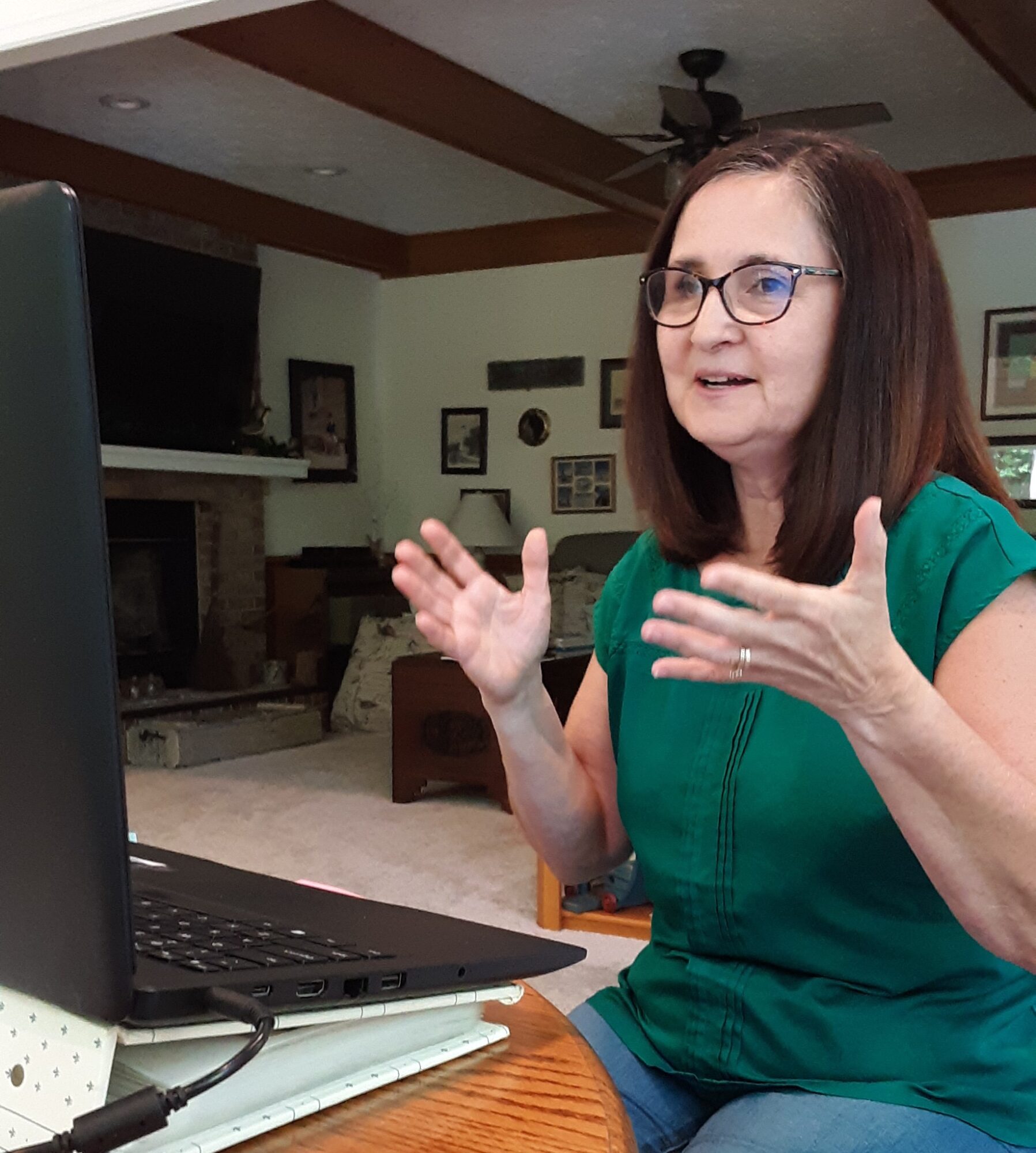Woman with glasses and shoulder-length hair gestures while sitting at a table with a laptop in a living room.