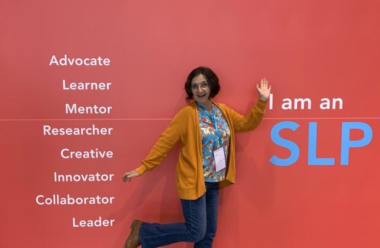 Woman standing next to a pink wall with words and large blue letters, waving hand, smiling, wearing glasses, a yellow cardigan, and a floral shirt.