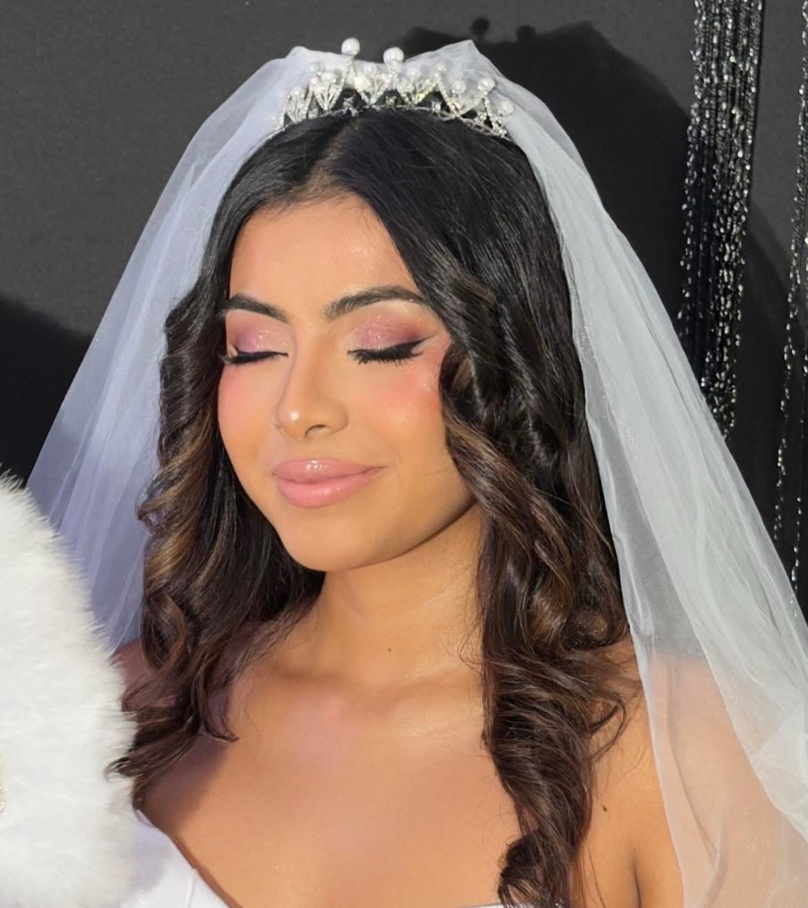 Young woman with closed eyes, wearing a wedding veil and tiara, smiling softly, with dark wavy hair and makeup.