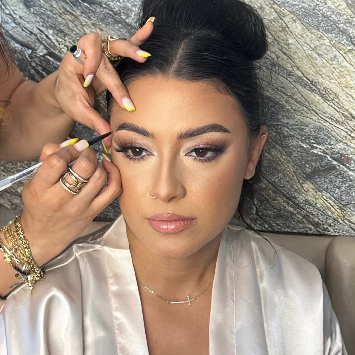 Woman getting makeup applied, lying against a textured stone background, with dark hair styled up, wearing a satin top and jewelry.