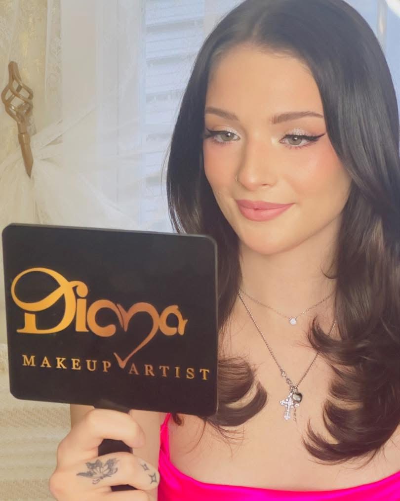 Young woman with dark hair and makeup holding a black sign with gold text, wearing a pink top and necklace.